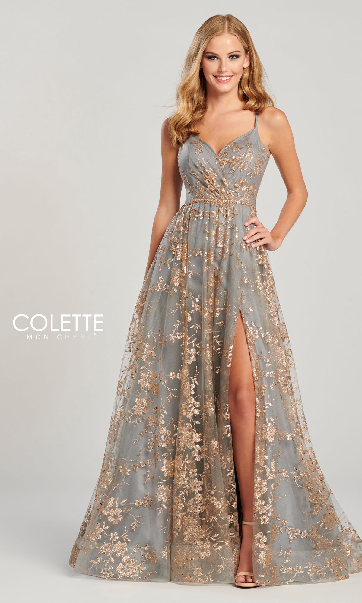 A woman poses in the Colette Gold and Pewter Long Prom Dress CL12006, a floor-length A-line gown with gold embroidery, glitter floral prints, deep V-neckline, thin straps, and a high leg slit against a plain background.